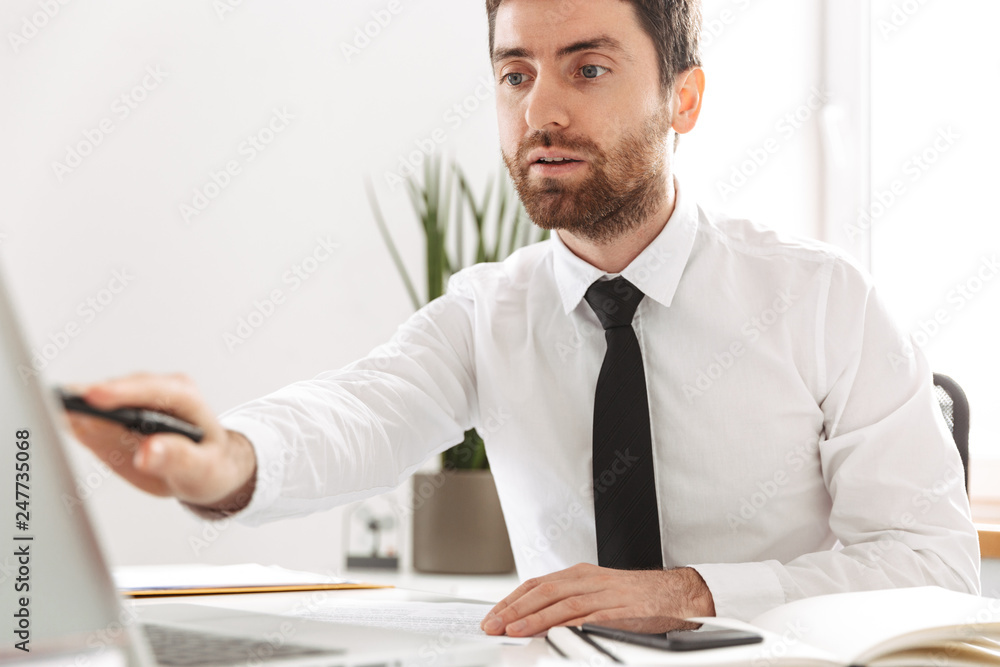Image of serious office worker 30s wearing white shirt working with laptop and notebook, in modern workplace Image of serious office worker 30s wearing white shirt working with laptop and notebook, in modern workplace