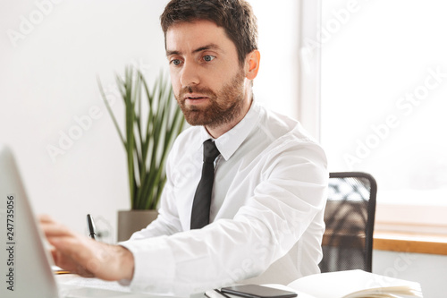 Image of european office worker 30s wearing white shirt working with laptop and notebook, in modern workplace © Drobot Dean