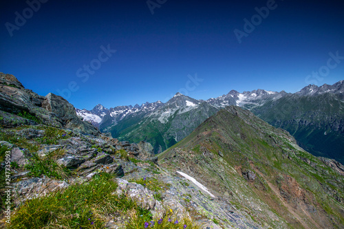 Mountain summit Dombai-Ulgen. In the background are glaciers and peaks of the Main Caucasus Range against the background of a piercing blue sky.