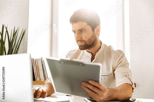 Portrait of serious businessman 30s wearing white shirt working with laptop and paper documents, while sitting in bright office