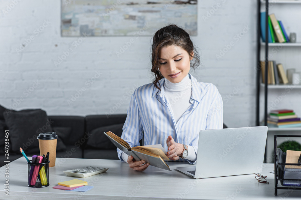 cheerful woman reading book near laptop in modern office cheerful woman reading book near laptop in modern office