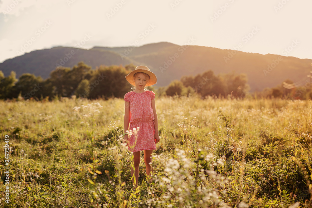 Cute blonde girl in straw hat and pink dress in the summer field at sunset