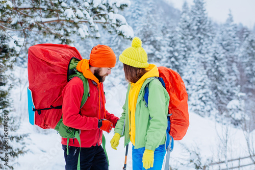 Couple hikers consulting during trekking in white winter woods at mountains. Young people walking on snowy trail with backpacks, healthy lifestyle adventure, camping, hiking trip concept.