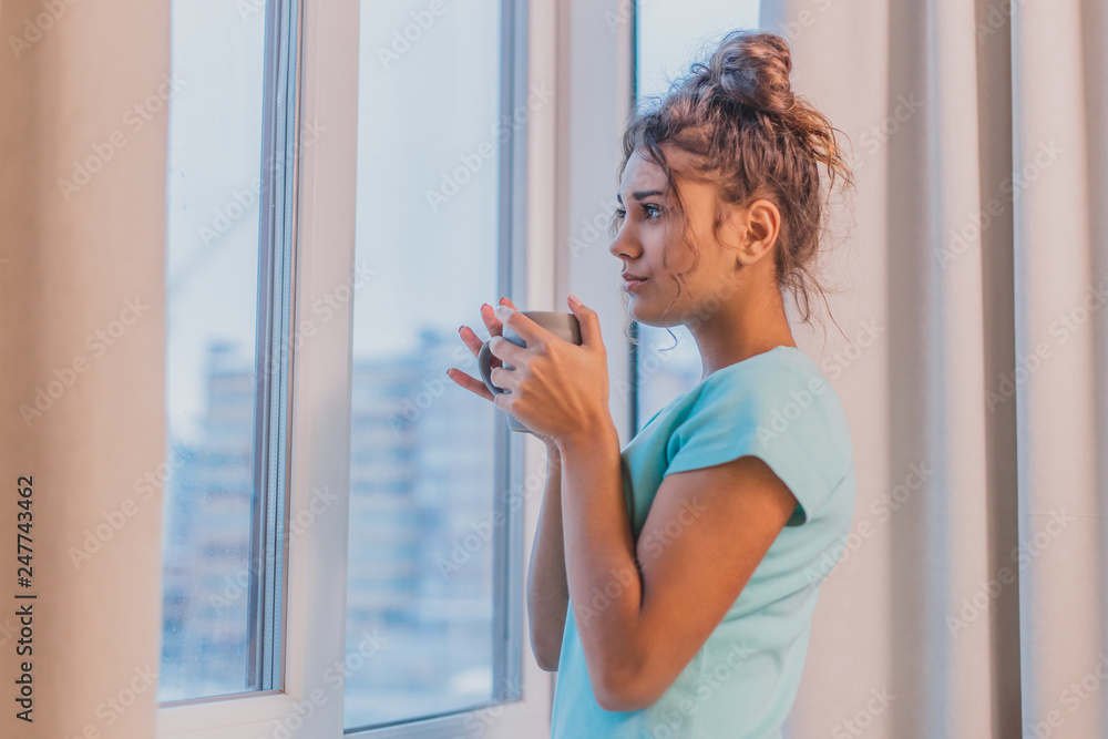Woman in the morning holding a cup of tea or coffee and looking at the sunrise standing near the window Woman in the morning holding a cup of tea or coffee and looking at the sunrise standing near the window