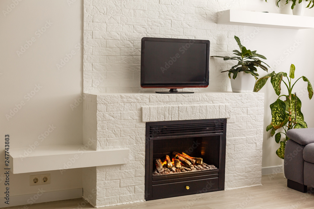 Room in a modern style. There is a TV on the white wall, white rack with a flower in vase, TV remote, decorations in a form black fireplace Room in a modern style. There is a TV on the white wall, white rack with a flower in vase, TV remote, decorations in a form black fireplace