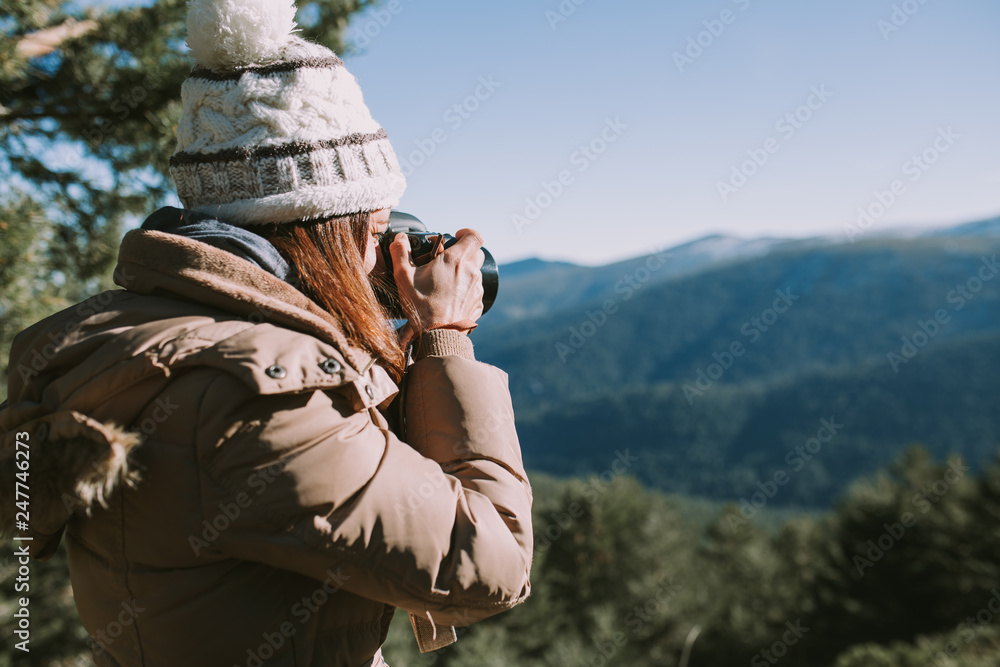 A brunette girl in a wool cap and brown coat takes a picture in the middle of the mountain. In the distance you can see snow-capped mountains and thick pine forests. A brunette girl in a wool cap and brown coat takes a picture in the middle of the mountain. In the distance you can see snow-capped mountains and thick pine forests.