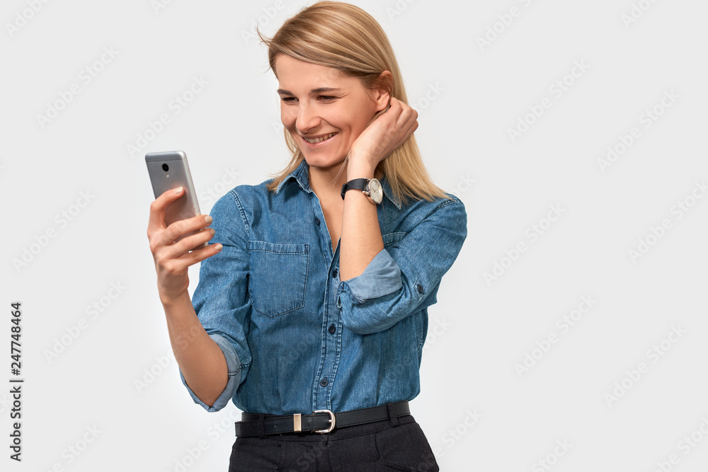 Portrait of cheerful blonde woman, holds modern smart phone, feeling happy to recieve good news, dressed in denim shirt, poses against white studio background with blank space for your advertisement Portrait of cheerful blonde woman, holds modern smart phone, feeling happy to recieve good news, dressed in denim shirt, poses against white studio background with blank space for your advertisement