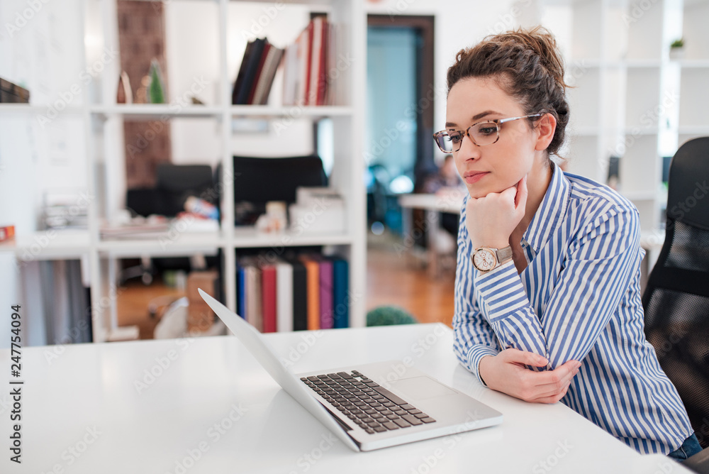 Young female manager in blouse with stripes looking at laptop at workplace, side view. Young female manager in blouse with stripes looking at laptop at workplace, side view.