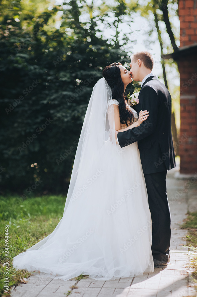 A young brunette bride in a white dress with a bouquet and a stylish groom in a black suit are hugging in a park with greenery. Portrait of lovers and smiling newlyweds. Wedding photography.