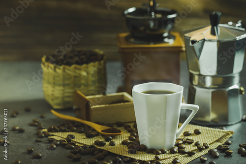 Brew black coffee in a white cup and morning lighting. Roasted coffee beans in a bamboo basket and wooden spoon. Vintage coffee grinder and pot. Concept of coffee time in morning or start the new day.