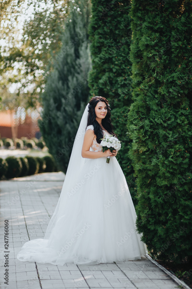 Cute bride in a white dress with a bouquet poses in the garden with greens and thujas. Portrait of a beautiful and smiling brunette. Wedding photography.