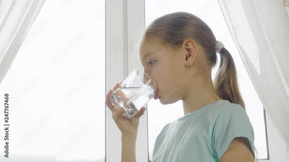 Girl teenager drinking clear water from glass on home kitchen on window background. Thirsty girl drinking fresh water from glass and looking to camera. Close up backlit natural light Girl teenager drinking clear water from glass on home kitchen on window background. Thirsty girl drinking fresh water from glass and looking to camera. Close up backlit natural light