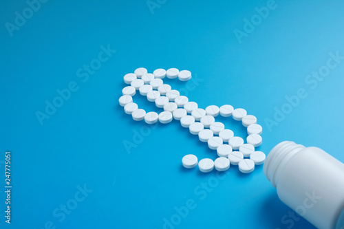 white pills spilling onto white table from white bottle into a dollar sign shape diagonally. Flat lay. Copy space.