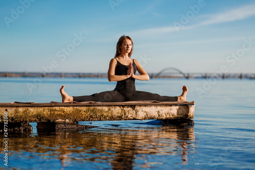 Young woman practicing yoga exercise at quiet wooden pier with city background. Sport and recreation in city rush