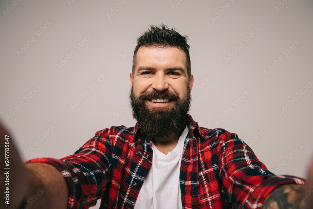 Fototapeta premium cheerful bearded man in checkered shirt taking selfie, isolated on grey
