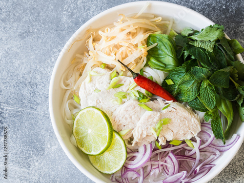 Traditional Vietnamese soup- pho ga in white bowl with chicken and rice noodles, mint and cilantro, green and red onion, chili, bean sprouts and lime on grey background. Asian food. Copy space