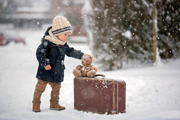 Baby playing with teddy in the snow, winter time. Little toddler boy in blue coat, holding suitcase and teddy bear, playing outdoors in winter park
