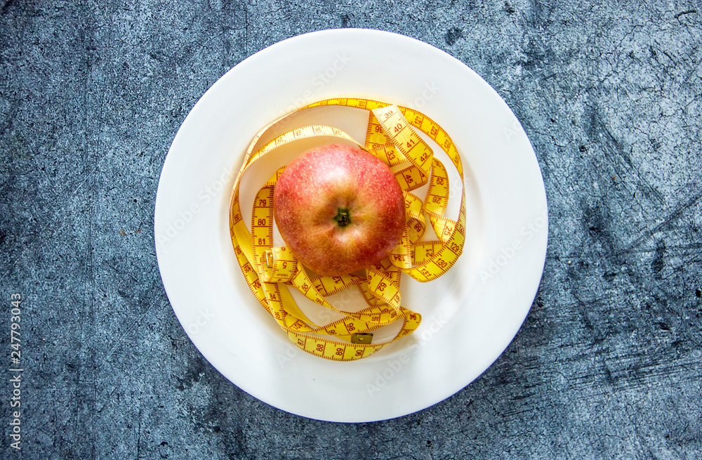 An apple on a white plate next to measuring tape and cutlery, a background about diets, a background about losing weight, the theme of vegetarianism An apple on a white plate next to measuring tape and cutlery, a background about diets, a background about losing weight, the theme of vegetarianism