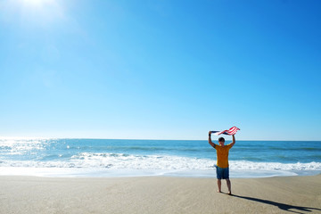 Young athlete running towards ocean holding american flag. Fit surfer man waving United States of America flag. 4th of July Independence day celebration concept. Back view, copy space.