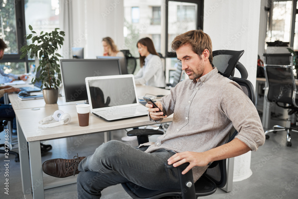 CEO with serious facila expression sitting in his chair and using smart phone. Office interior, in background employees working. CEO with serious facila expression sitting in his chair and using smart phone. Office interior, in background employees working.