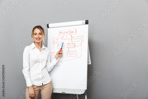 Image of smiling businesswoman in formal wear standing and making presentation using flipchart in the office, isolated over gray background