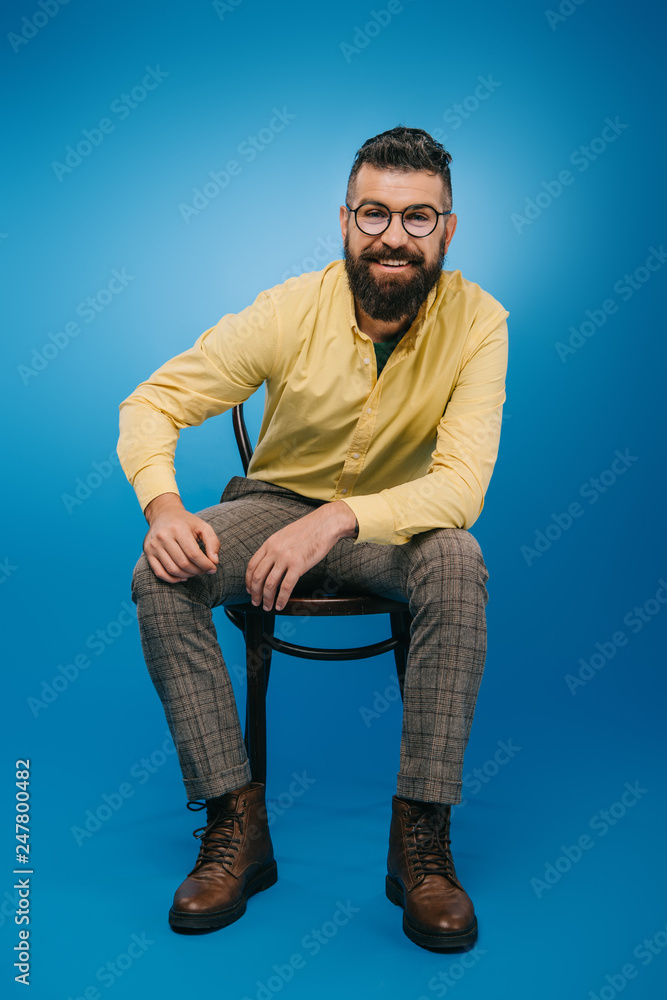 smiling bearded man in eyeglasses sitting on chair isolated on blue smiling bearded man in eyeglasses sitting on chair isolated on blue