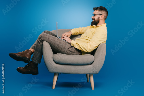 bearded businessman using laptop on armchair on blue