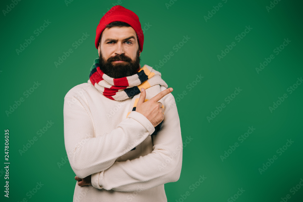 sick bearded man in hat and scarf pointing aside isolated on green sick bearded man in hat and scarf pointing aside isolated on green