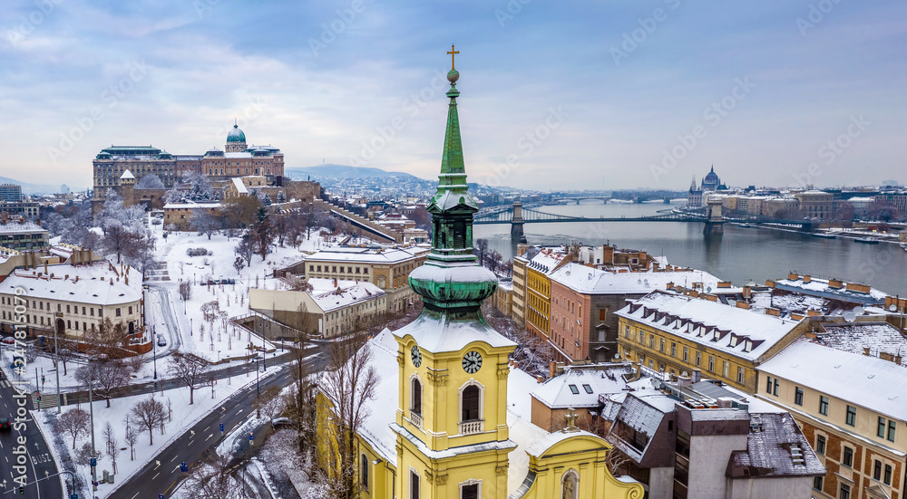 Fototapeta premium Budapest, Hungary - Catholic church with snowy Buda district, Buda Castle Royal Palace, Varkert Bazaar, Szechenyi Chain Bridge and Parliament at background on a snowy winter morning