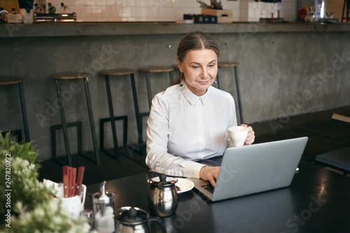 Candid shot of fashionable smiling busy middle aged female entrepreneur checking email on her generic laptop computer while having cappuccino during break, sitting in cozy coffee house interior