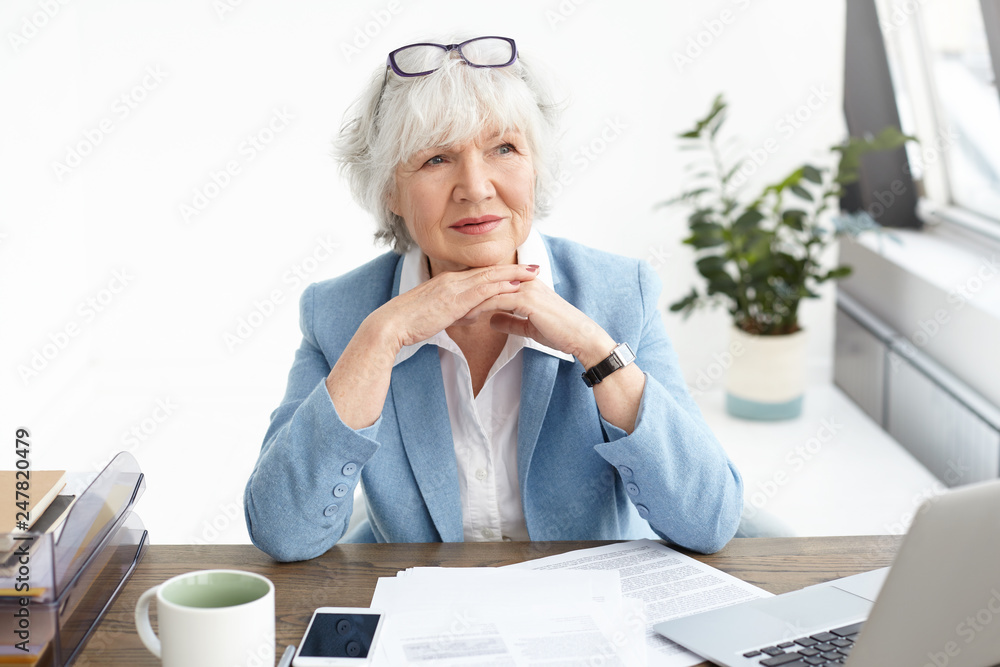Business, people, electronic gadgets and modern technology concept. Indoor shot of experienced senior Caucasian female CEO in formal wear having pensive look while working at desk with laptop Business, people, electronic gadgets and modern technology concept. Indoor shot of experienced senior Caucasian female CEO in formal wear having pensive look while working at desk with laptop