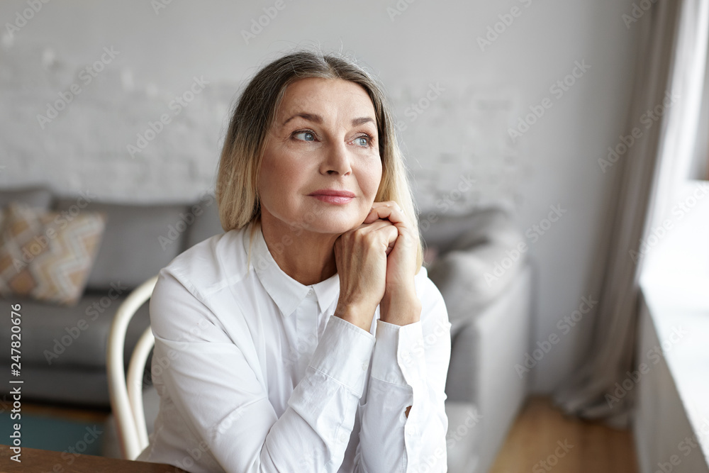 Indoor portrait of beautiful 50 year old Caucasian woman with thick loose hair sitting by the window in living room having dreamy pensive look, daydreaming or thinking about something pleasant Indoor portrait of beautiful 50 year old Caucasian woman with thick loose hair sitting by the window in living room having dreamy pensive look, daydreaming or thinking about something pleasant
