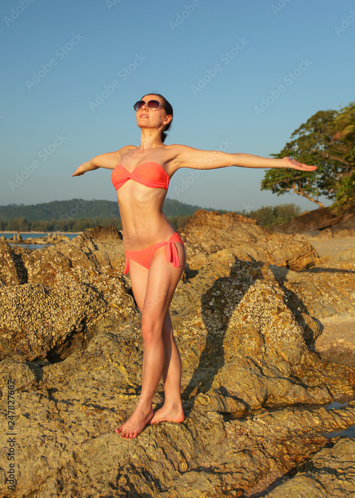 Young slim woman in bikini swim wear and sunglasses, standing on the rocks  near beach, arms spread, facing to sun, as if gathering energy. Stock Photo  | Adobe Stock