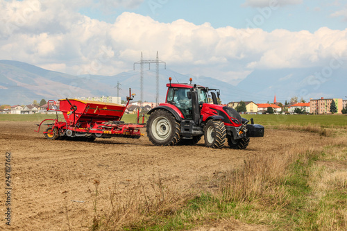 Red tractor sowing  pulling seeder trailer behind  on dry field  some houses and mountains in background.