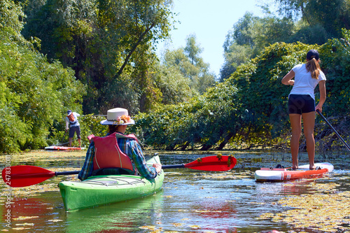 Teen girl on stand-up paddle board SUP and woman in green kayak paddling in wilderness river overgrown duckweed near thickets of trees and wildgrapes at sunny summer day photo