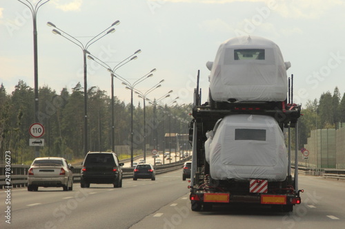 Heavy car carrier truck transports new cars in protective covers on semi-trailer on suburban asphalt highway road on summer day against forest and blue sky - logistics business in Europe, delivery