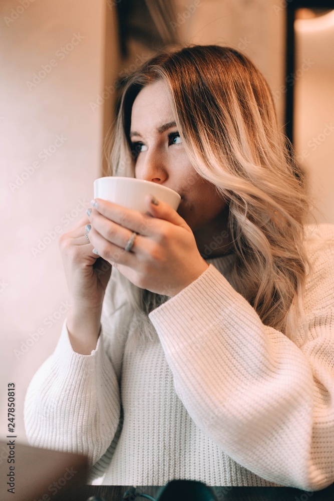 blonde girl in a cozy warm cafe, in a white knitted sweater drinking coffee.