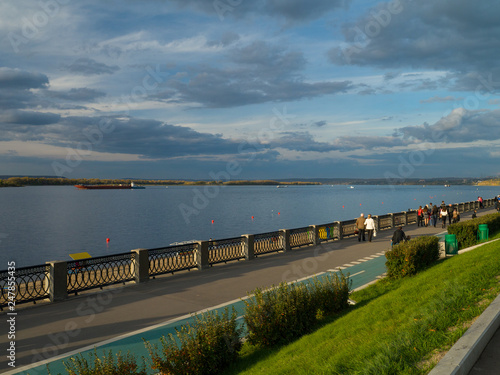 View of the city embankment with an openwork pig-iron protection, a green grass and the passing people in solar autumn evening against the background of the blue sky with clouds.