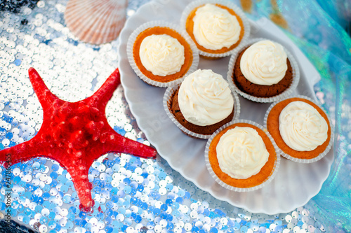 Vanilla cupcackes with white creamcheese on seashell shape plate and big red sea star near it. Silver fabric on the background photo