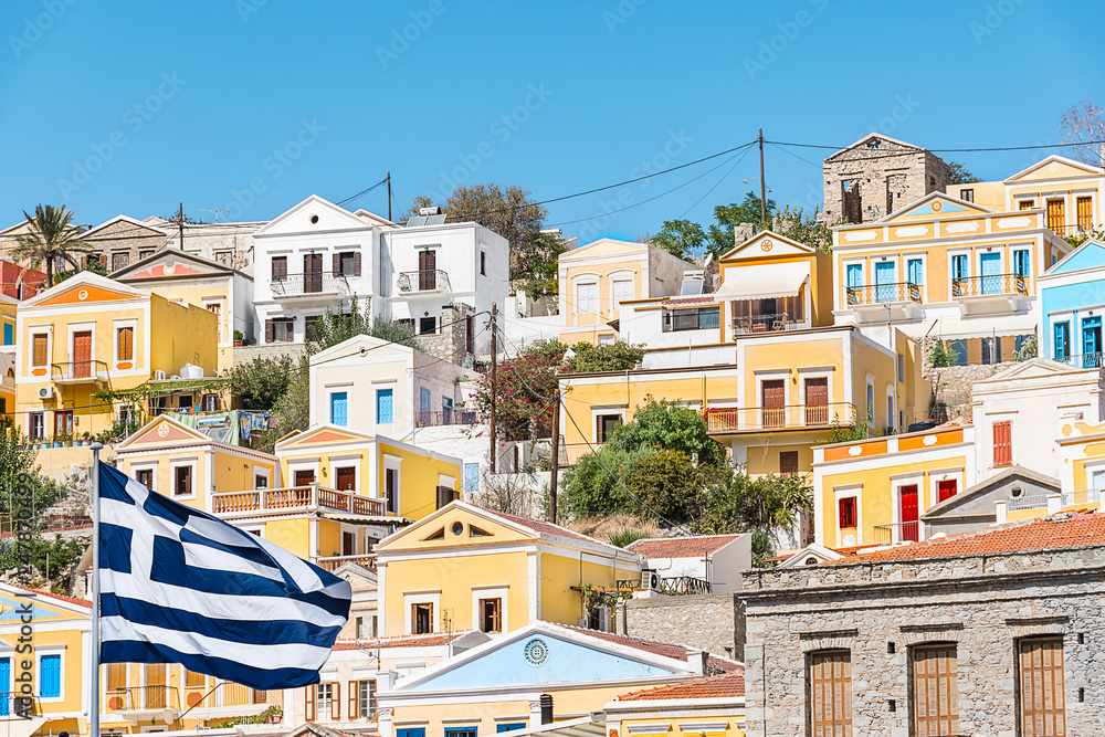 Greek flag and colorful neoclassical houses in harbor town of Symi (Symi Island, Greece)