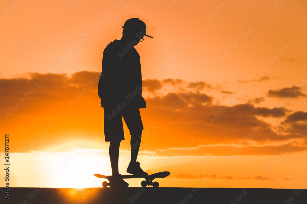 Silhouette of young boy plays with skateboard at sunset near Backlight of a skater with hat standing in a orange sky with golden reflections on the ocean. Teenager playing with a foot on the board