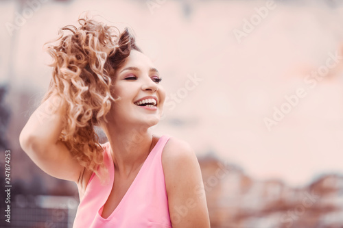 close-up portrait of a young beautiful sexy girl with curly hair in a pink feather dress smiling lifestyle on a street in sunny day