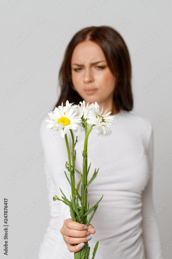 Studio portrait of allergic brunette female in white top holding a bouquet of flowers, having an allergic reaction to chamomile, selective focus.Allergy, medical, seasonal flowers concept Studio portrait of allergic brunette female in white top holding a bouquet of flowers, having an allergic reaction to chamomile, selective focus.Allergy, medical, seasonal flowers concept