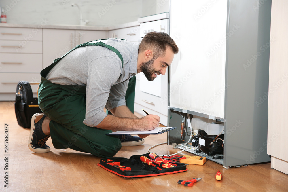 Male technician with clipboard examining refrigerator in kitchen Male technician with clipboard examining refrigerator in kitchen