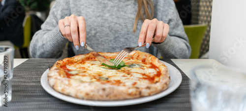 Woman eats with knife and fork a pizza Margherita with mozzarella tomatoes and basil. Neapolitan pizza from wood-burning stove. lunch in an Italian restaurant. Table near to a large window.