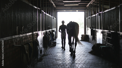Quadro su tela Young jockey walking with a horse out of a stable
