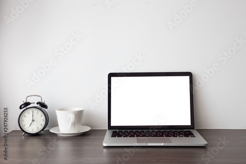 wood office table with blank screen on laptop, notebook and hot coffee cup, retro black clock on white cement background.