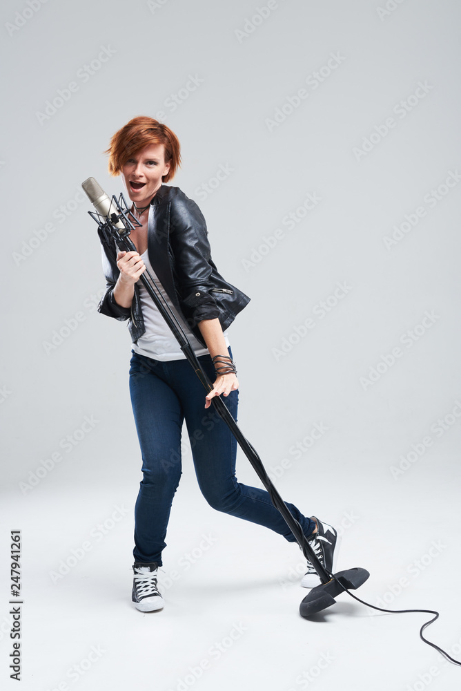 Fototapeta premium Full-length portrait of rock singer wearing leather jacket and keeping static mic, sings a song loudly on grey background. Concept of rock music and rave