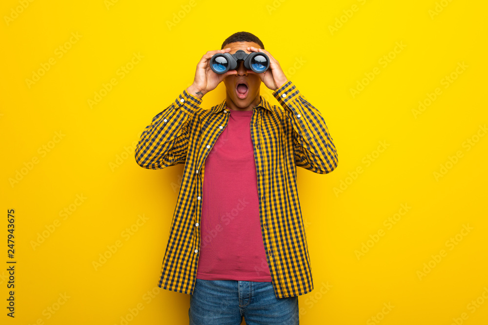 Fototapeta premium Young afro american man on yellow background and looking in the distance with binoculars