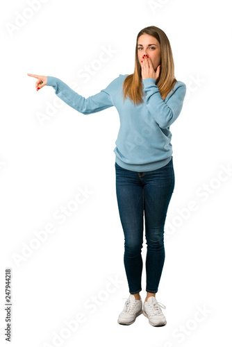 Full body of Blonde woman with blue shirt pointing finger to the side with a surprised face on white background
